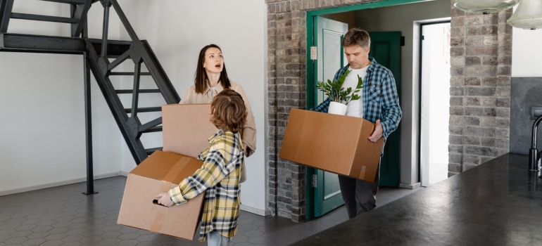 a family of three walking into a new home while carrying cardboard boxes