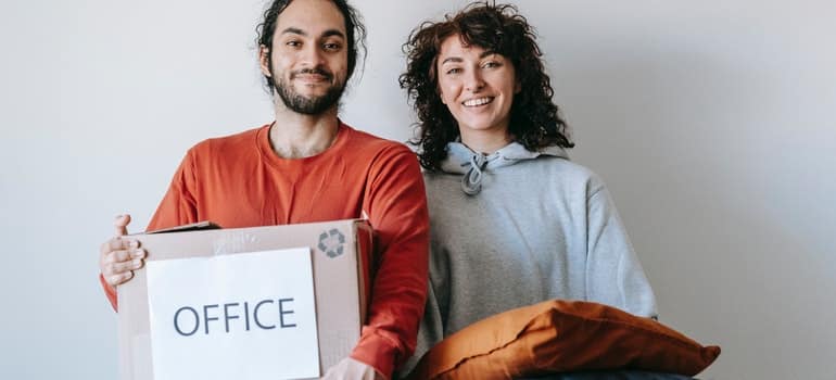 A man holding a whilte box with a word office printed on it and a woman holding a pillow, while thinking how to engage employees during an office move;
